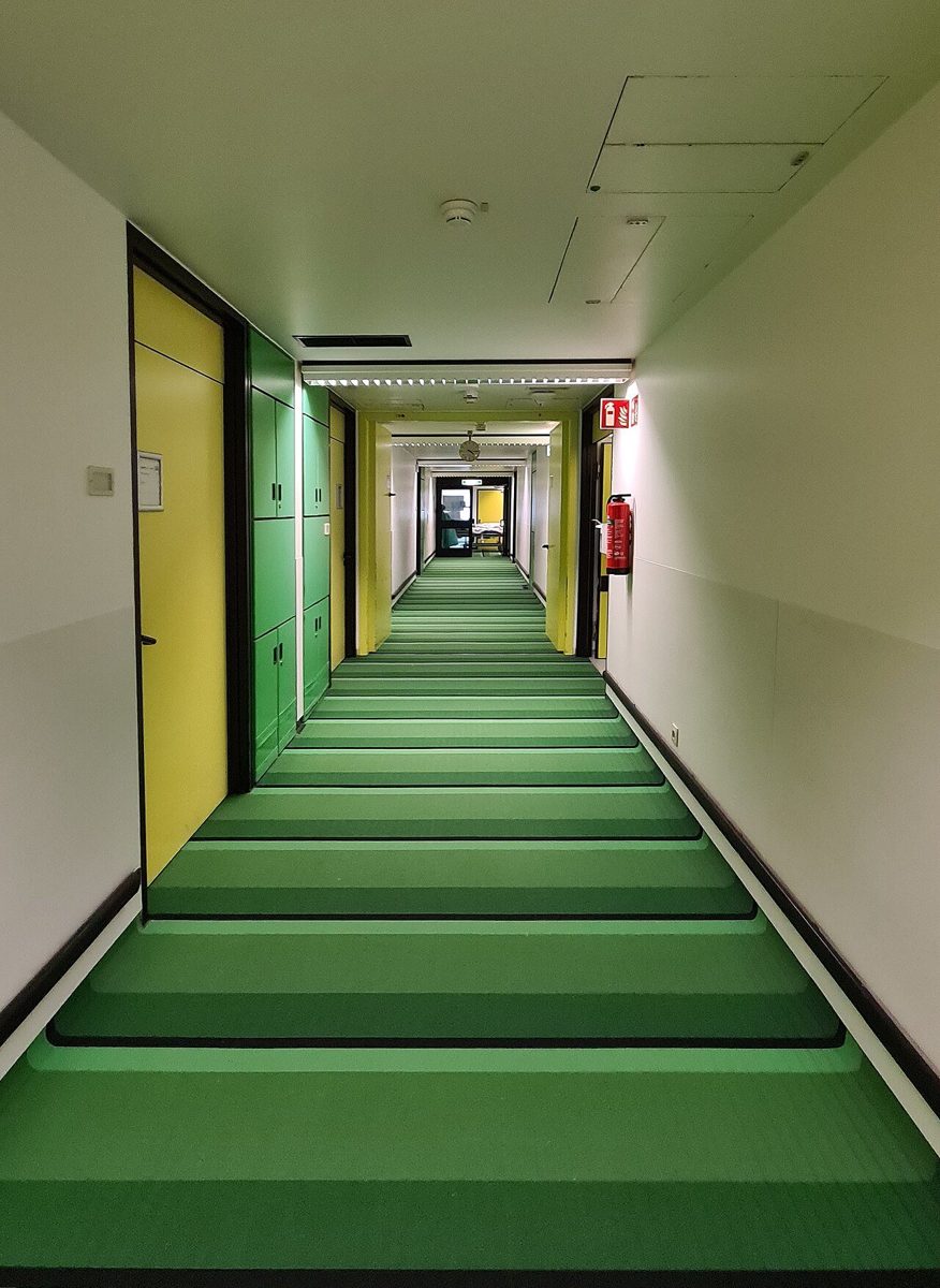 Long hospital corridor with a green resilient floor laid in bands, yellow doors and green cabinets