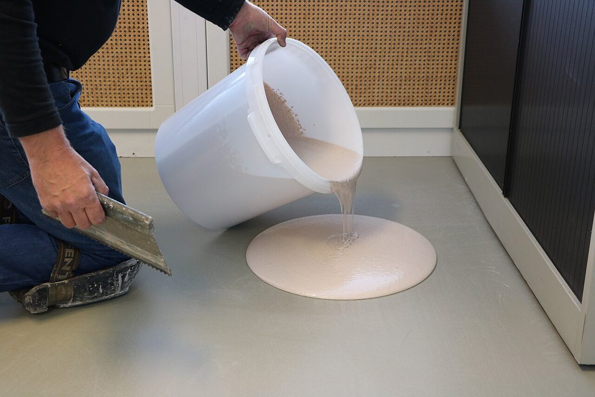 Hands pouring a liquid floor compound from a bucket onto a grey floor, notched trowel in the other hand