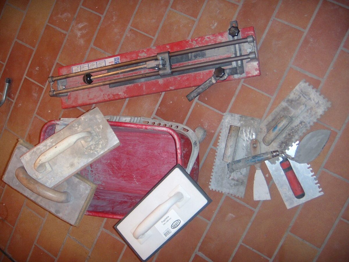 Tiling tools on a terracotta floor: a tile cutter, grout floats, notched trowels, a putty knife and a tub