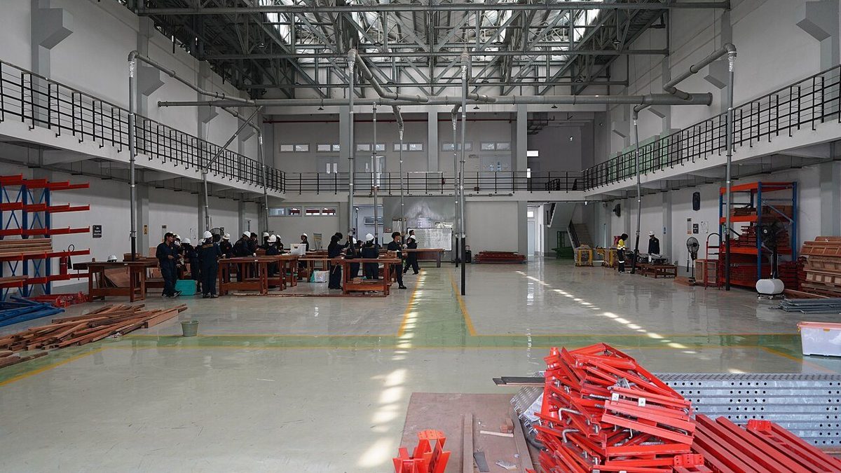 Large vocational workshop hall with timber workbenches, red steel parts and a group of trainees at the far end
