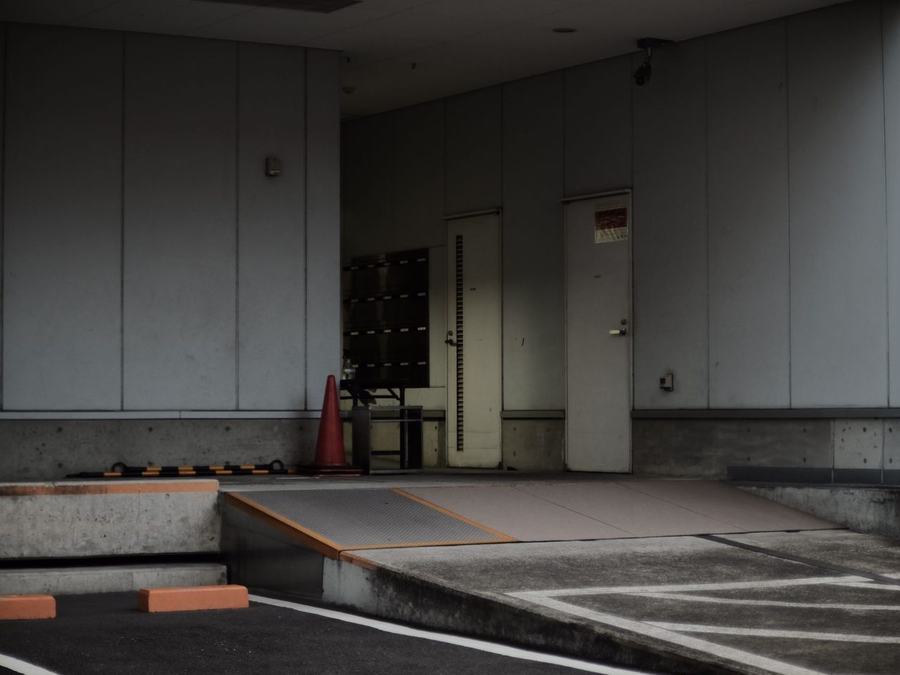 Concrete building entrance with a sloped driveway leading to a raised walkway, painted parking lines, doors and orange traffic cones near the wall.
