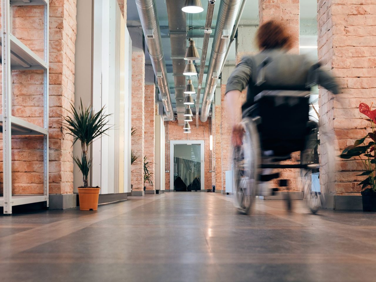 Blurred person in a wheelchair rolling down a long office hallway with a smooth gray floor, exposed brick columns, shelving and potted plants.