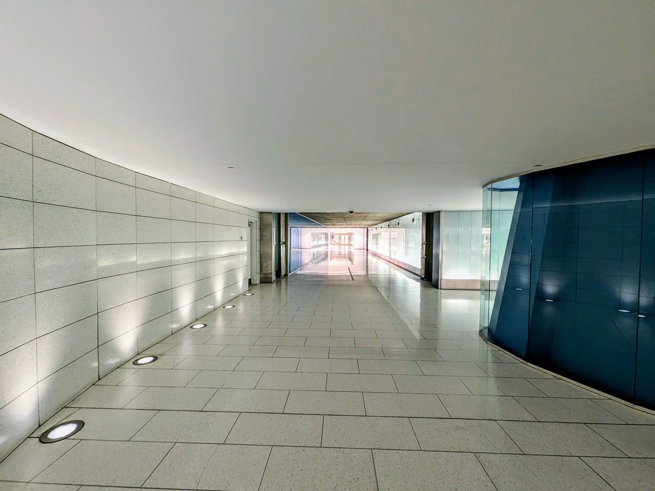 Long curved corridor with large gray floor tiles, round lights set into the floor along a white wall, and blue frosted glass panels.