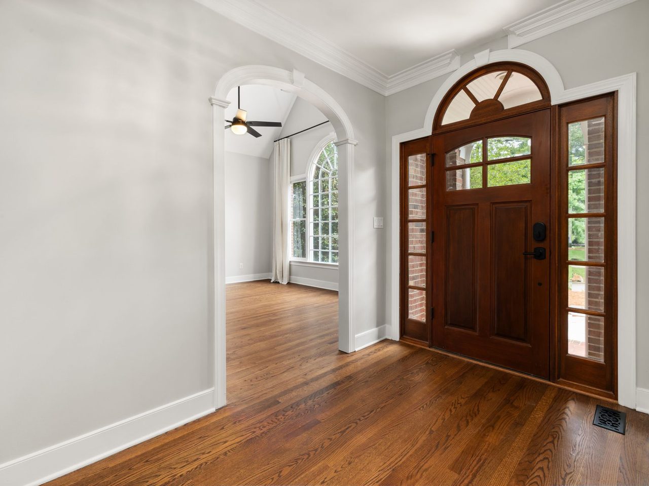 Empty foyer with hardwood floor leading to an arched wooden front door with glass sidelights, a metal floor vent and white walls.