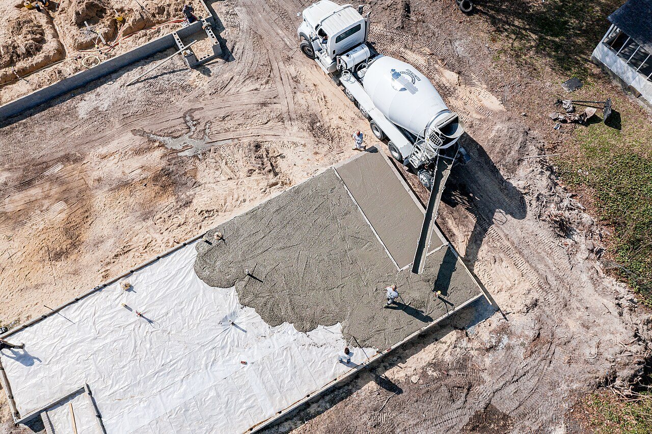 Aerial view of concrete spreading over white vapor barrier sheeting inside slab forms, a mixer truck alongside