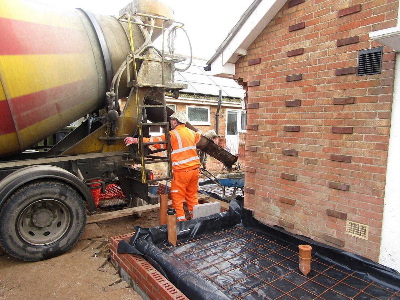 Black damp-proof membrane and steel mesh laid in a formed area beside a brick wall as a mixer truck unloads