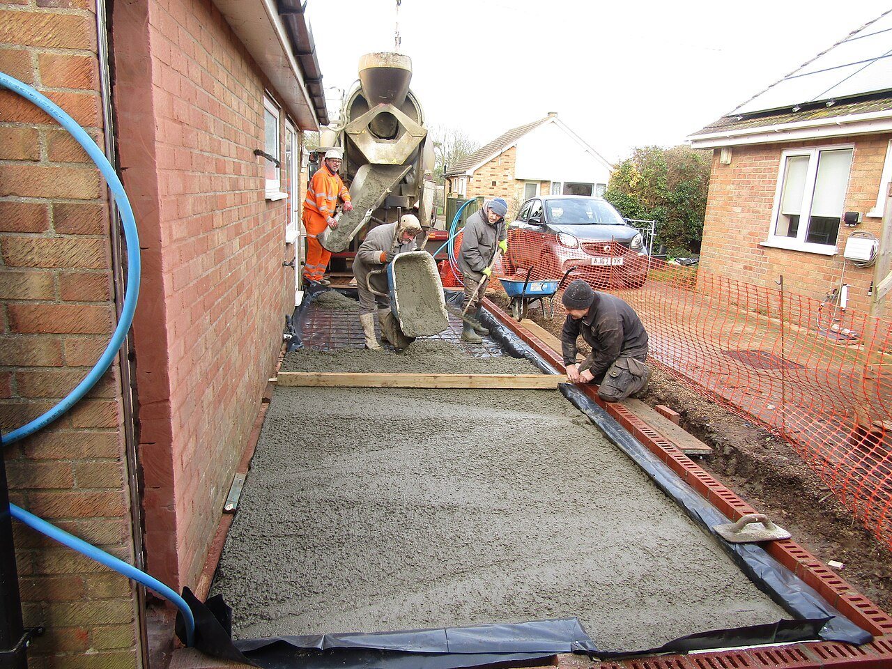 Crew spreading ready-mix concrete over a black membrane for a narrow slab beside a brick house