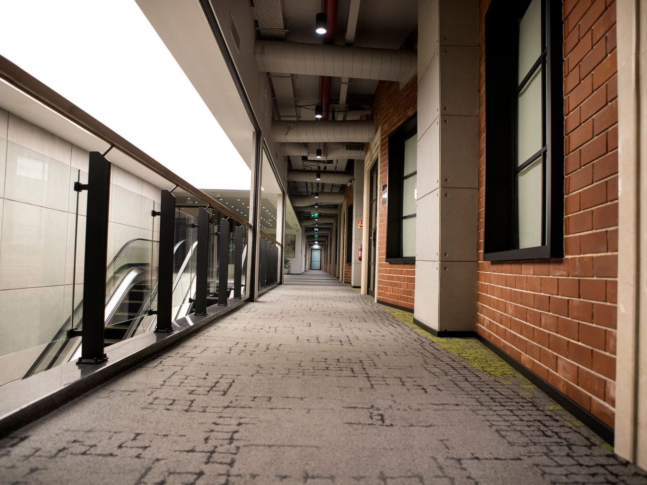 Long indoor hallway with gray patterned carpet, a red brick wall on one side and glass railings and an escalator on the other