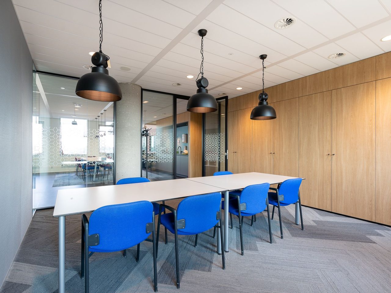 Office meeting room with gray carpet, a long white table, blue chairs, three black pendant lights, wood paneled wall and glass wall