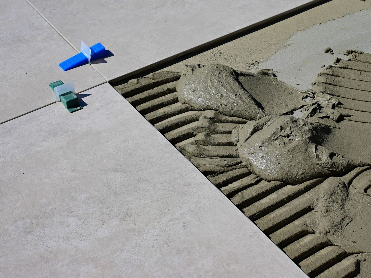 Close-up of gray mortar spread in notched-trowel ridges beside a laid gray floor tile, with blue and green plastic tile spacers at the joint.