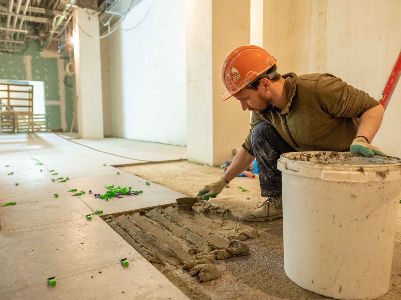 Worker in orange hard hat and gloves kneels beside a white bucket, spreading mortar and setting tiles on a floor in a gutted interior.
