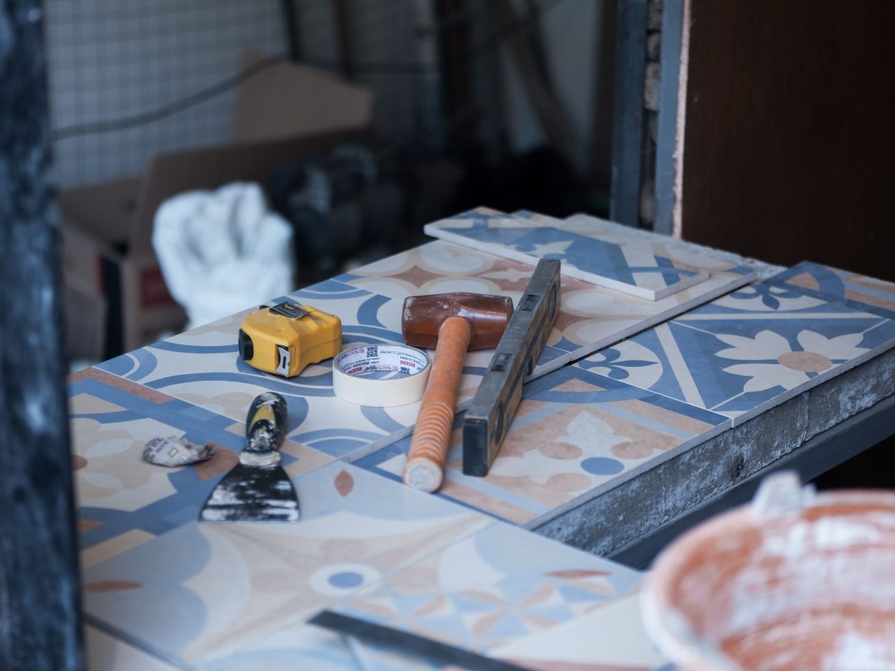 Blue and white patterned tiles on a work surface with a trowel, yellow tape measure, level, white gloves and a bucket on a construction site.