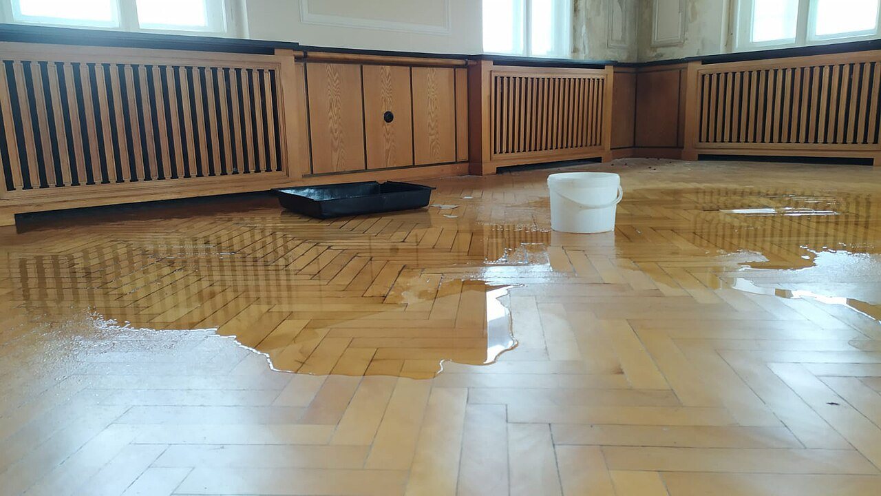 Water pooled across an oak herringbone floor in a large room, with a bucket and a paint tray