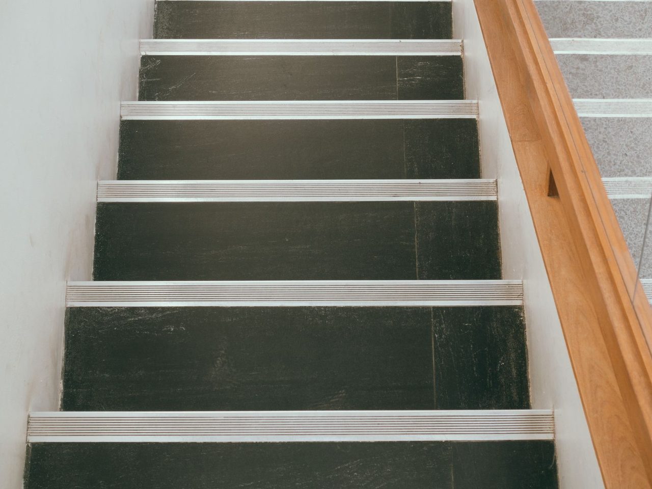 Staircase with dark treads and light metal nosing strips beside a wooden handrail