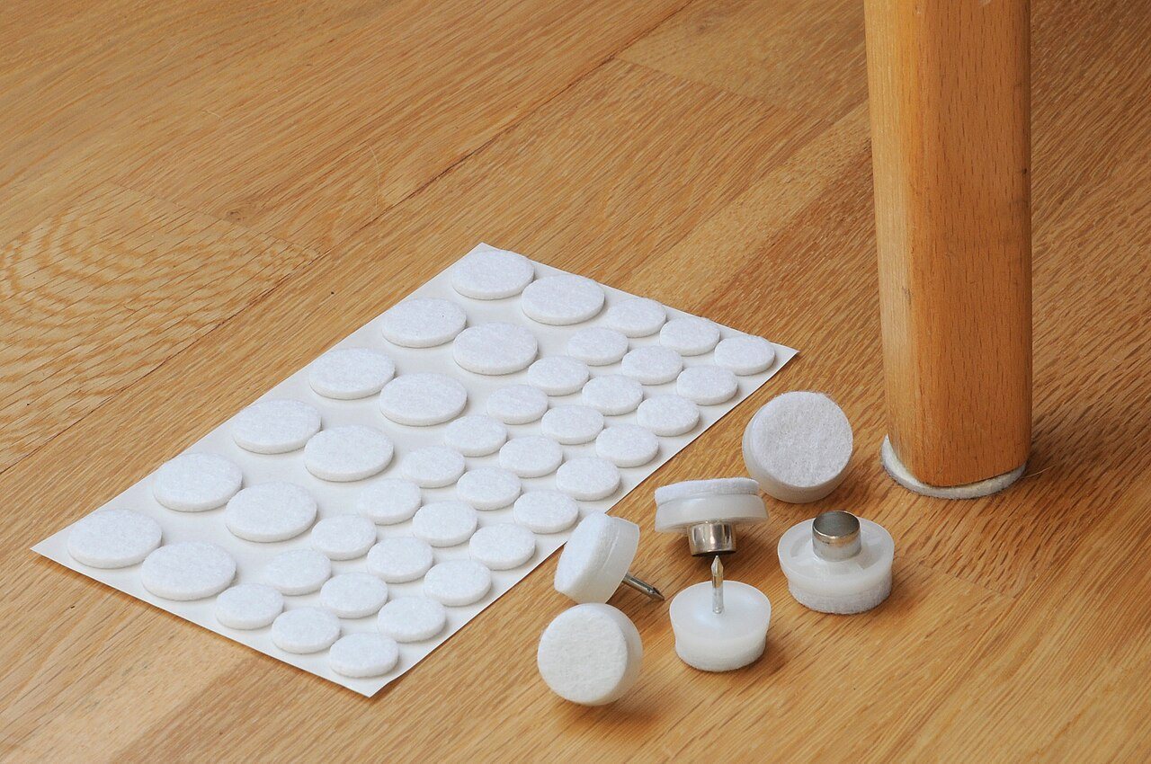 Sheet of white felt furniture pads and several glides next to a chair leg on a wooden floor