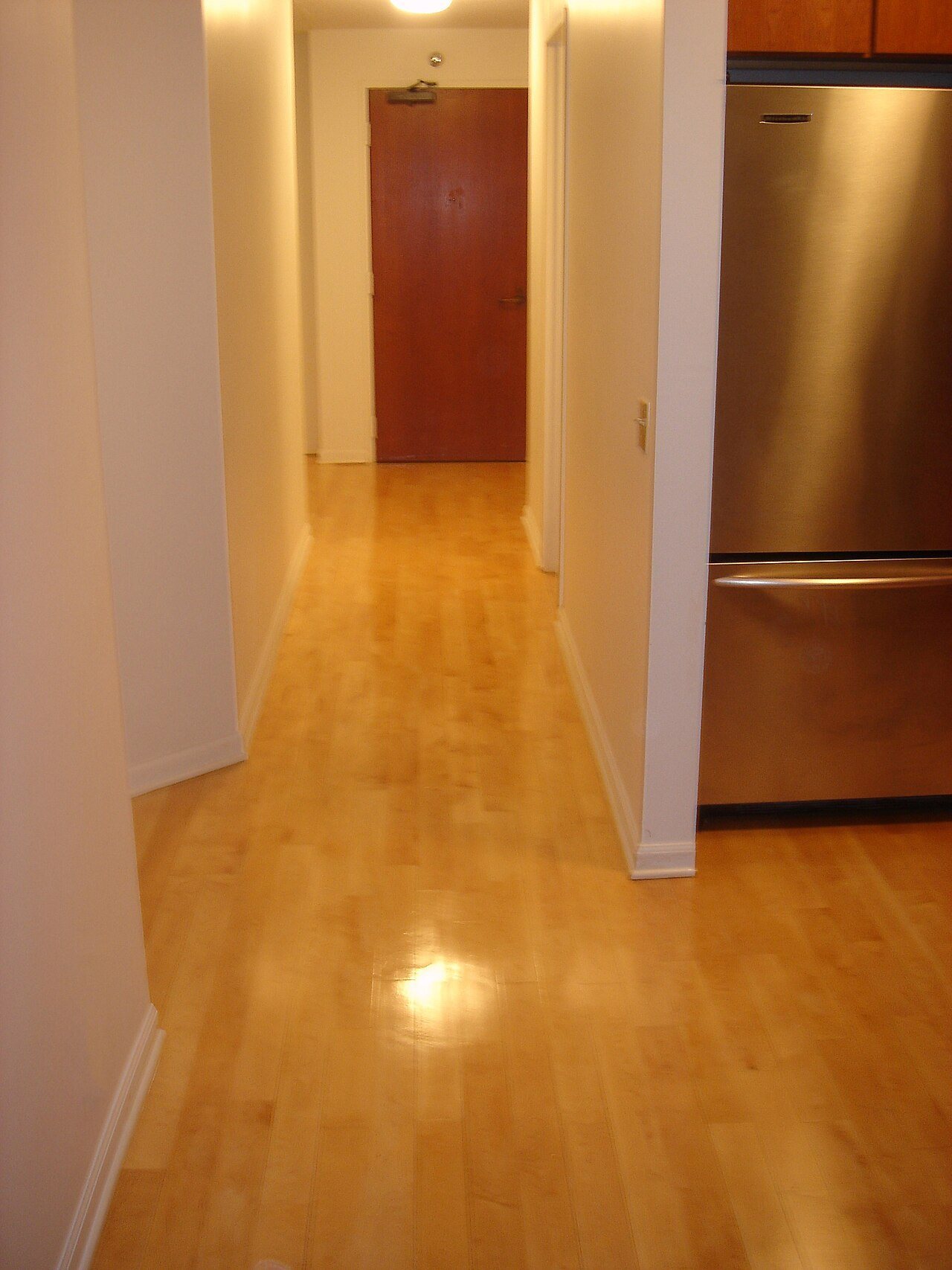Narrow apartment hallway with a light maple strip floor running past a stainless steel refrigerator