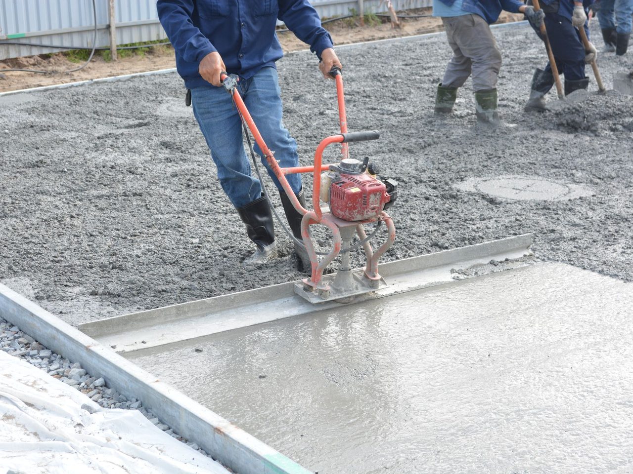Worker in jeans pulling a red motorized screed along a long metal bar over wet gray concrete, with other workers raking concrete in the background.