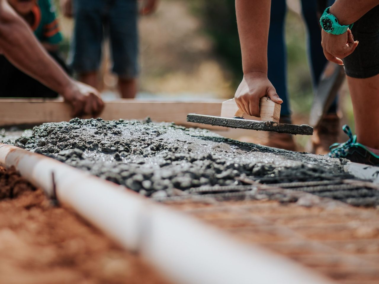 Two workers pulling a straight board across freshly placed concrete inside wooden forms, viewed close to the surface of the wet slab.