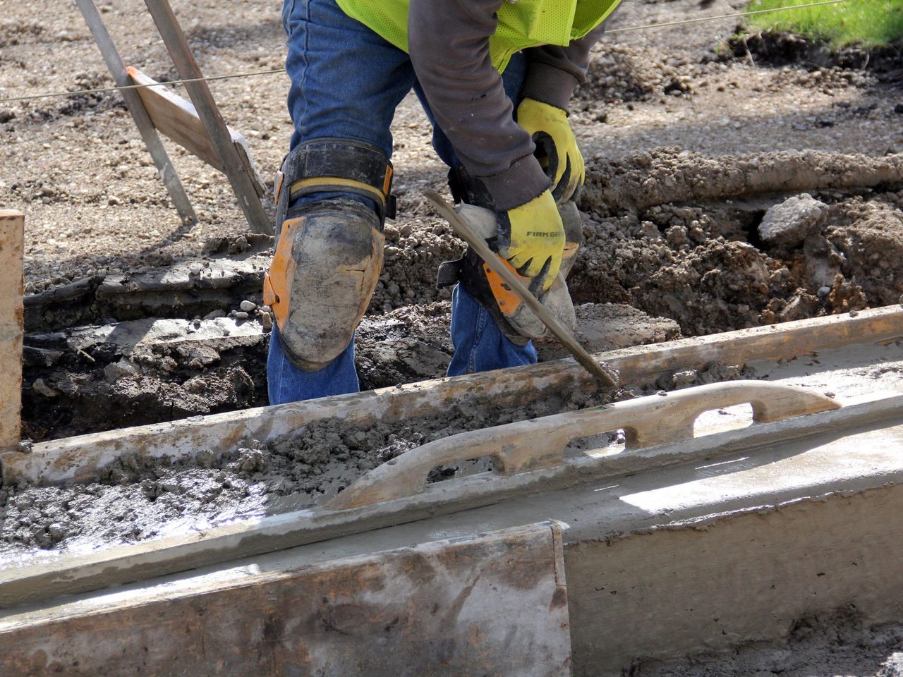 Kneeling worker in yellow gloves and knee pads using a hand trowel on a strip of wet concrete next to wooden form boards outdoors.