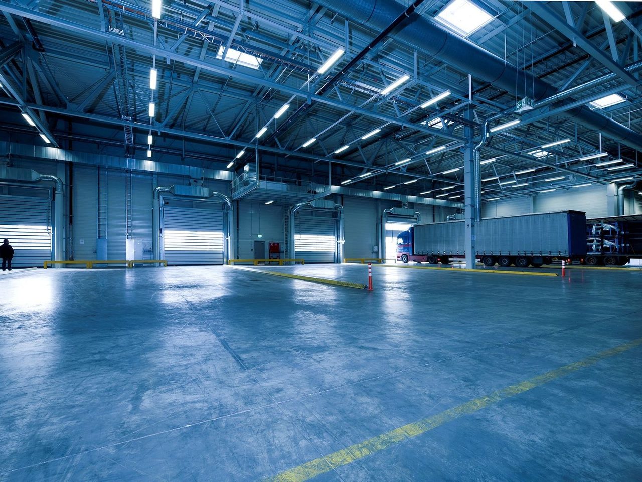 Wide interior of an empty warehouse with a smooth concrete floor, ceiling light rows, two people in the distance and trucks at loading doors.