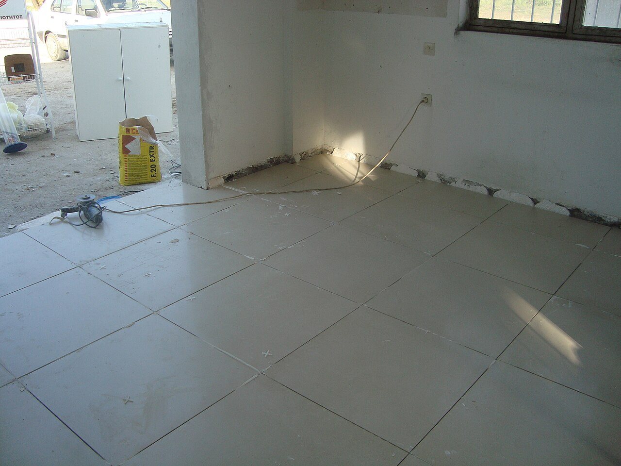 Freshly laid large off-white floor tiles in an unfinished room, with an angle grinder and adhesive bag nearby