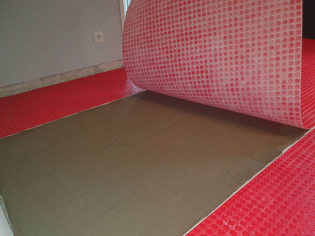 Red dimpled membrane rolled back from a bed of gray thinset combed with notched trowel ridges
