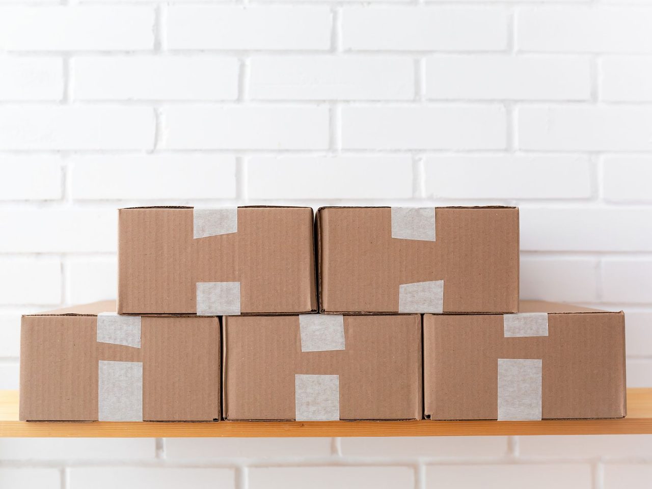 Taped cardboard cartons stacked on a wooden shelf against a white brick wall