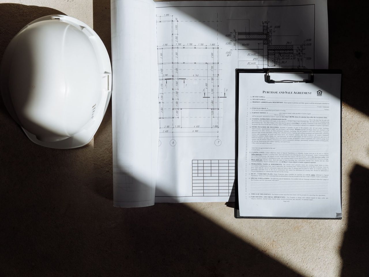 Top view of construction documents on a clipboard beside a white hard hat