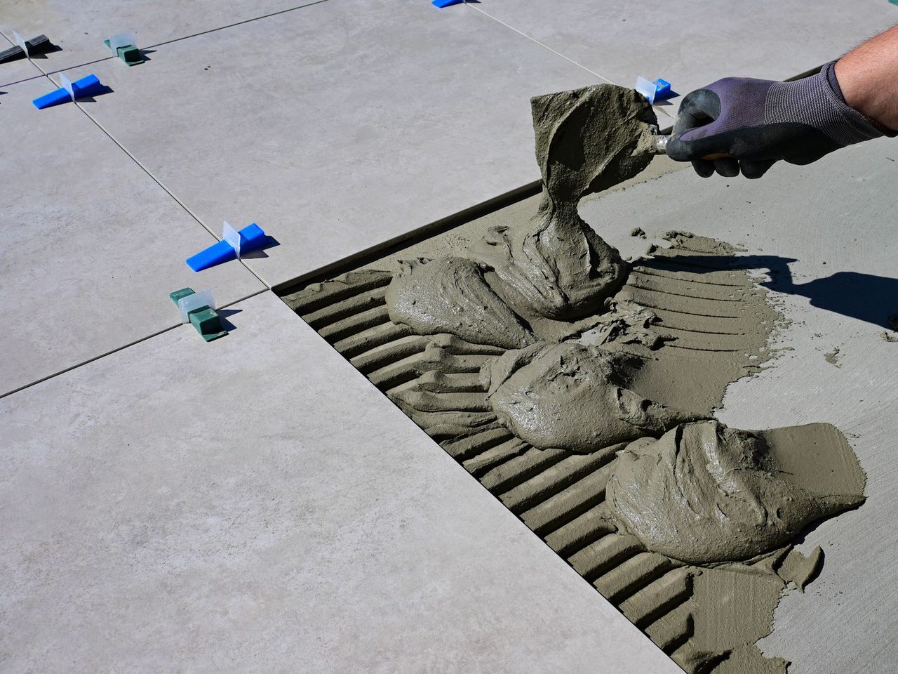 Gloved hand spreading mortar in notched ridges beside a set floor tile
