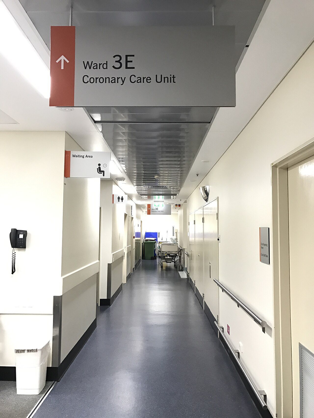 Hospital corridor with blue sheet vinyl, dark wall base, handrails and a Coronary Care Unit sign