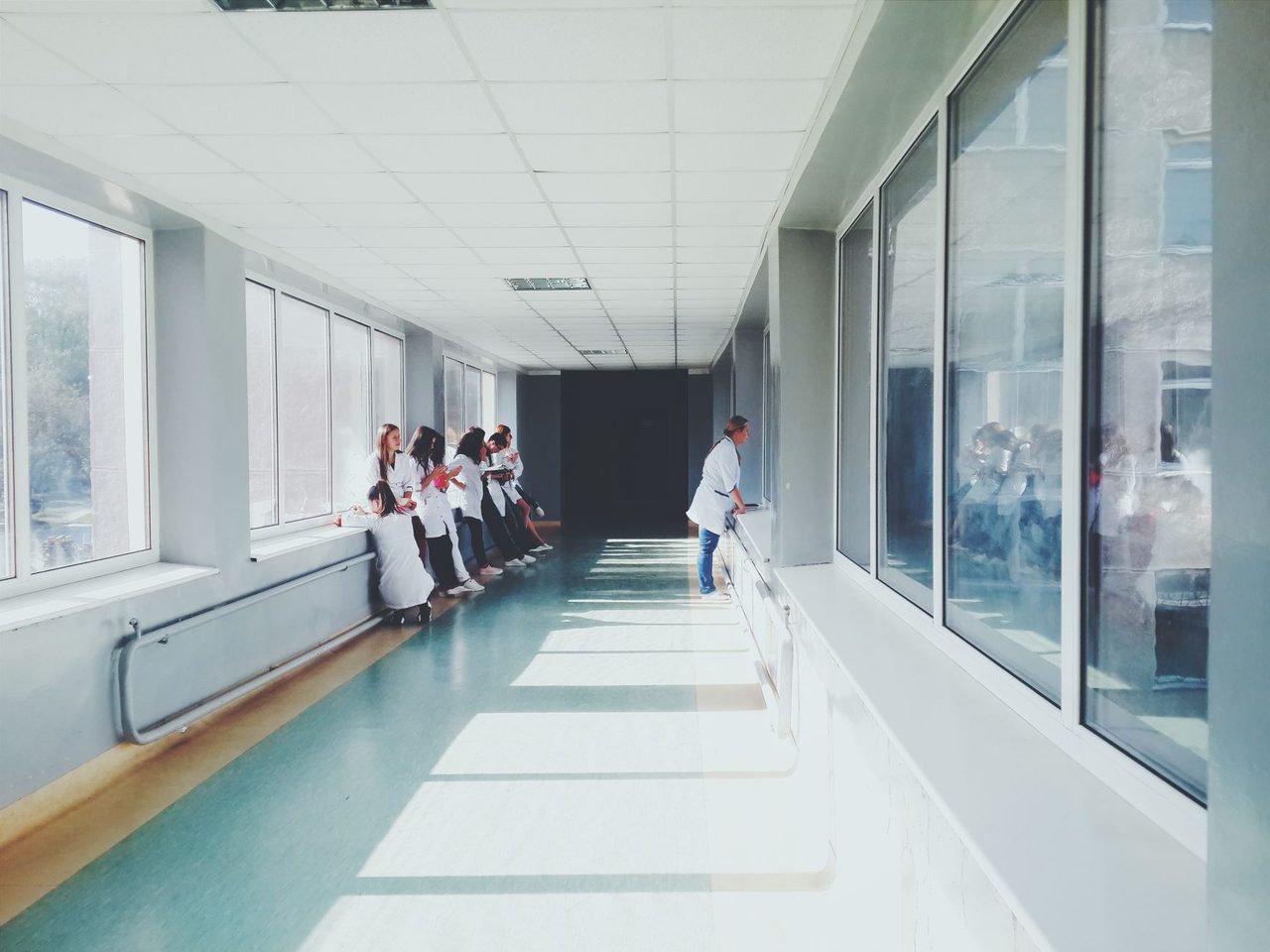 Long hospital hallway with a glossy teal floor, windows along both sides, a wooden bench and staff in white coats in the distance.