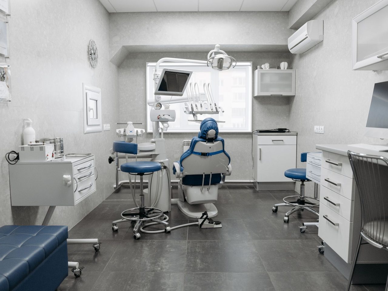 Dental clinic room with a blue and white dental chair, rolling stools, white cabinets, an overhead lamp and a gray floor.