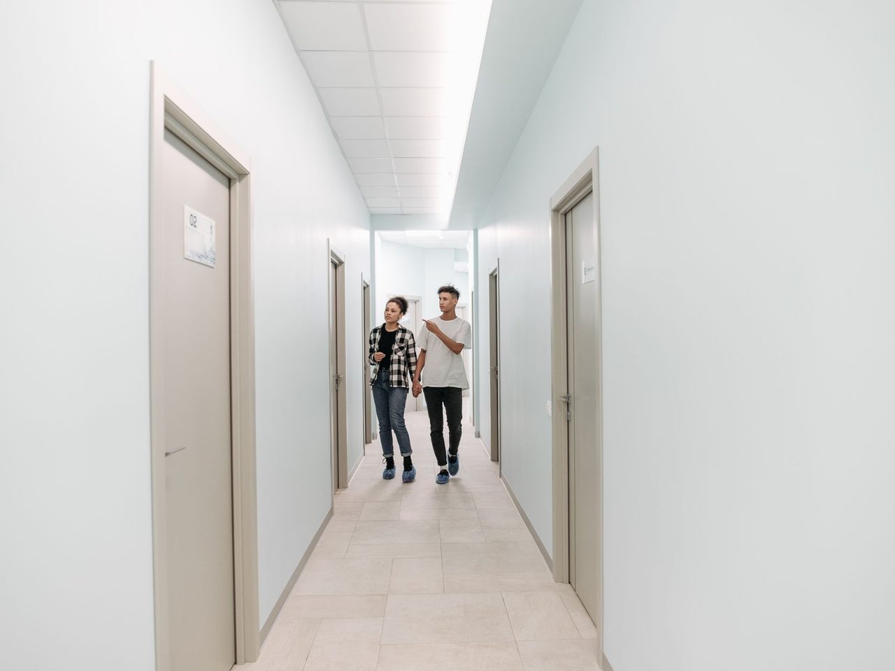 Two people walking down a bright white medical clinic corridor with a pale floor, recessed ceiling lights and gray doors on each side.