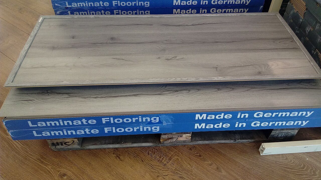 Grey oak-look laminate plank resting on blue cartons printed Laminate Flooring, Made in Germany