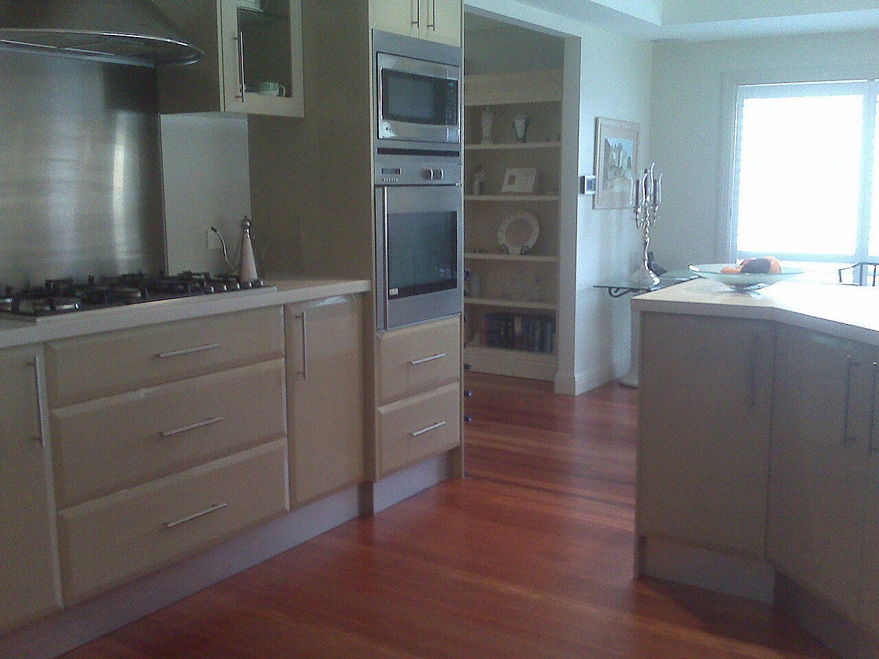 Kitchen with a reddish hardwood floor between a cabinet run with built-in ovens and an island