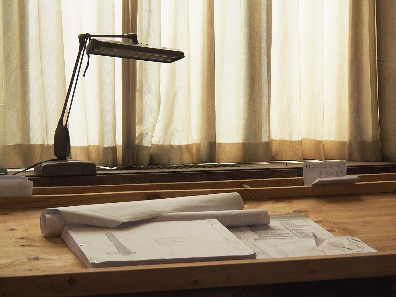 Rolled construction drawings and a bound plan set on a wooden desk under a drafting lamp by a curtained window