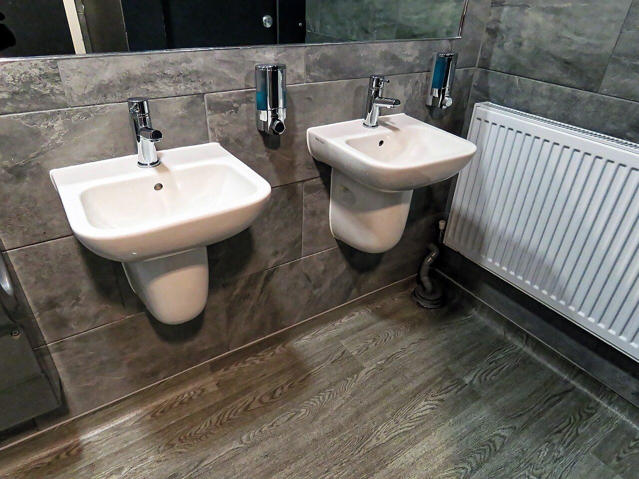Two wall-hung washbasins above a grey wood-look plank floor in a tiled washroom with a radiator