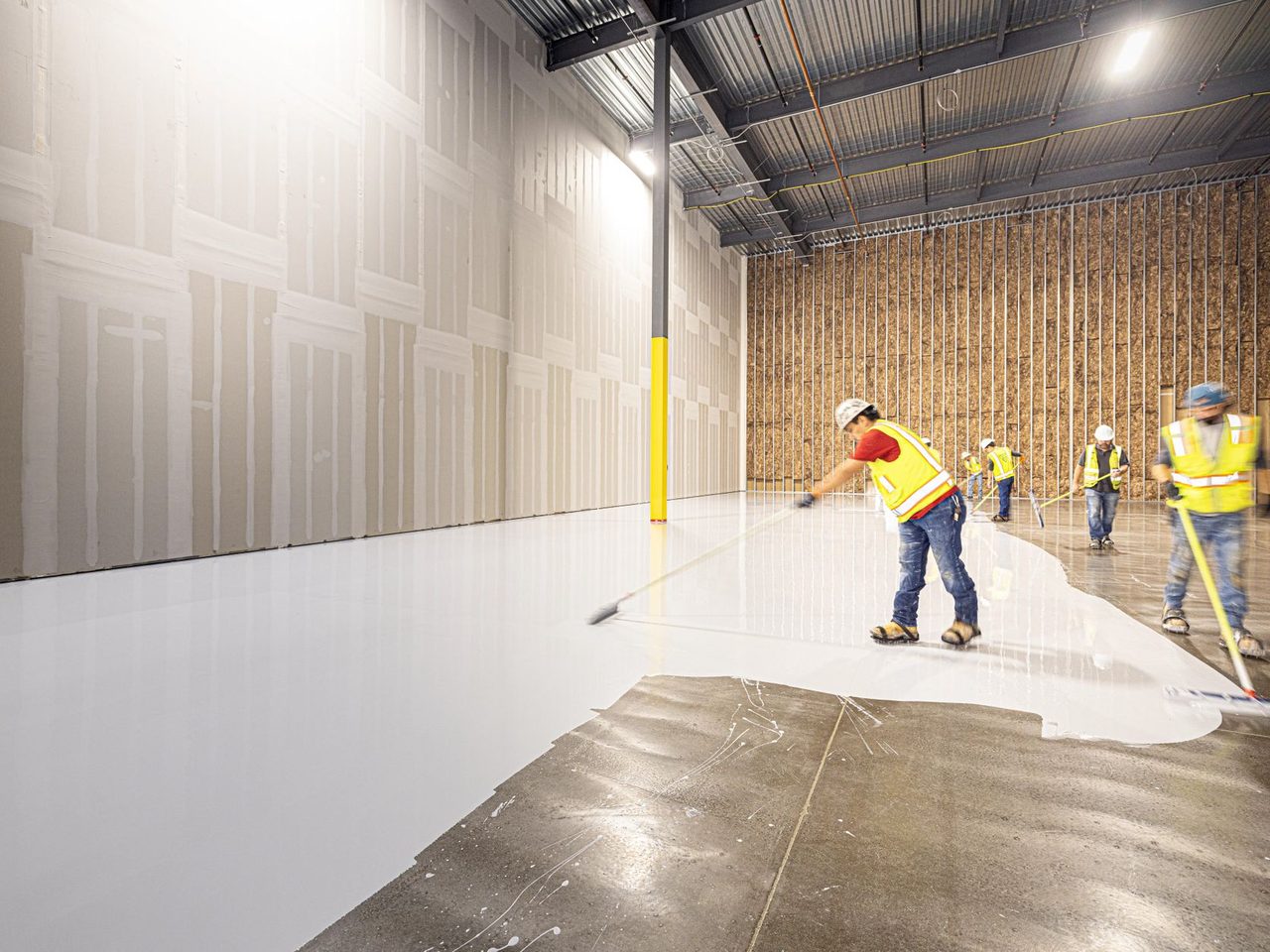 Workers in hard hats and safety vests spreading a fresh concrete floor inside a large building