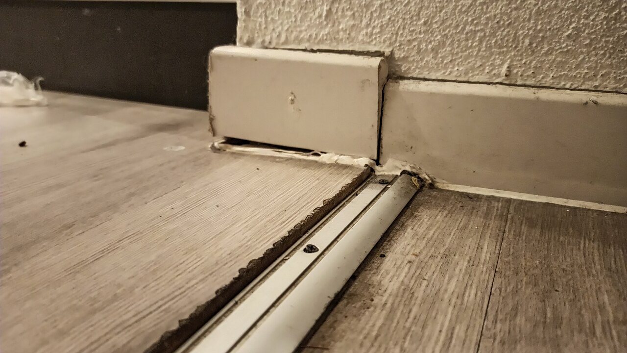 A screwed-down metal transition strip between two wood-look plank floors, beside a baseboard with a visible gap at its foot