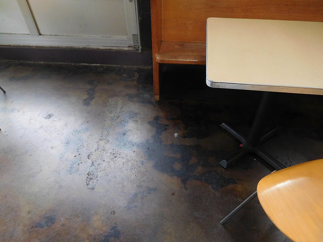 Patchy concrete floor with dark blotches and pale worn areas beside a table and chair