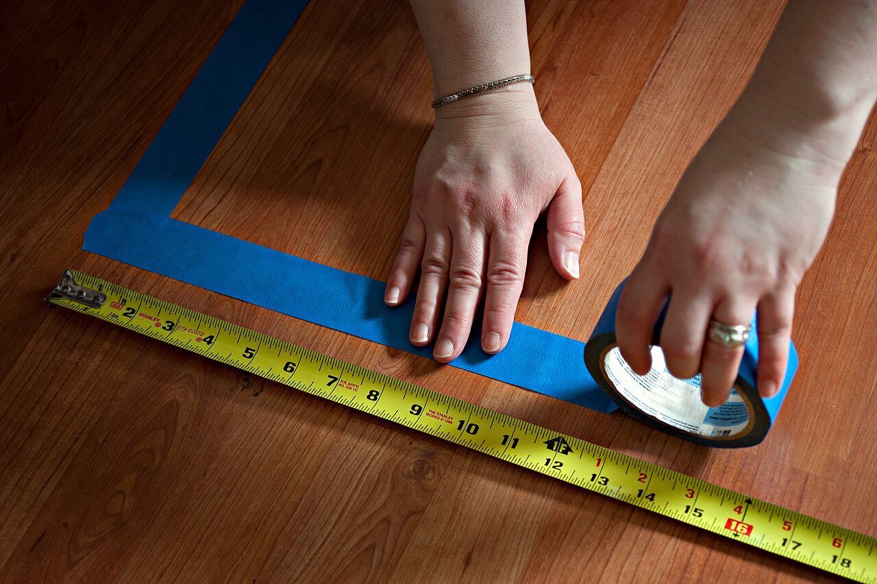 Two hands laying blue painter's tape in a rectangle on a wood floor beside an extended yellow tape measure