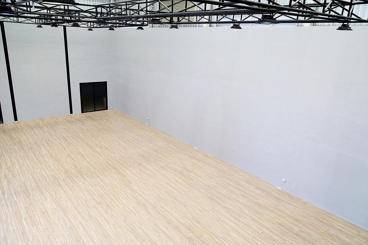 Large empty hall with a light wood-look vinyl plank floor, white walls and black steel roof trusses
