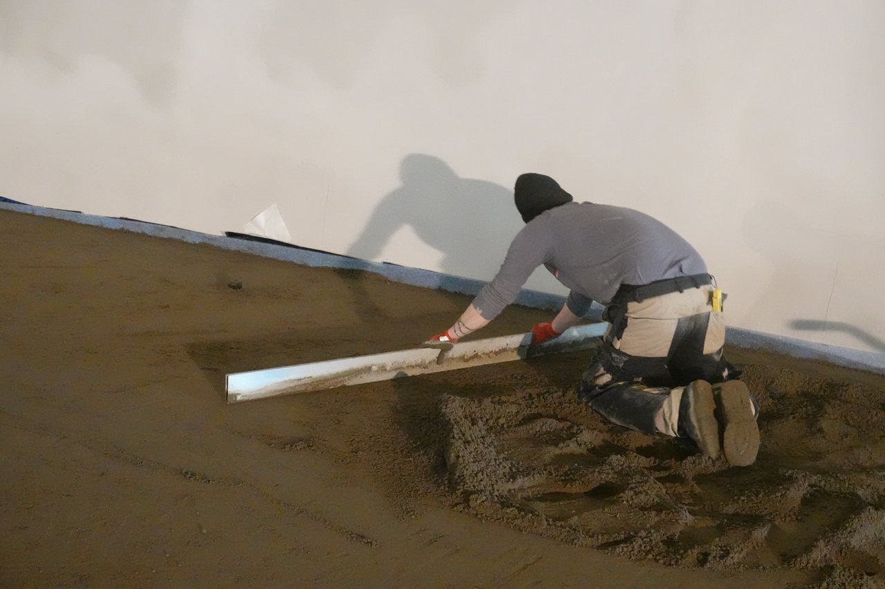Kneeling worker levelling semi-dry screed with a long aluminum straightedge beside a wall