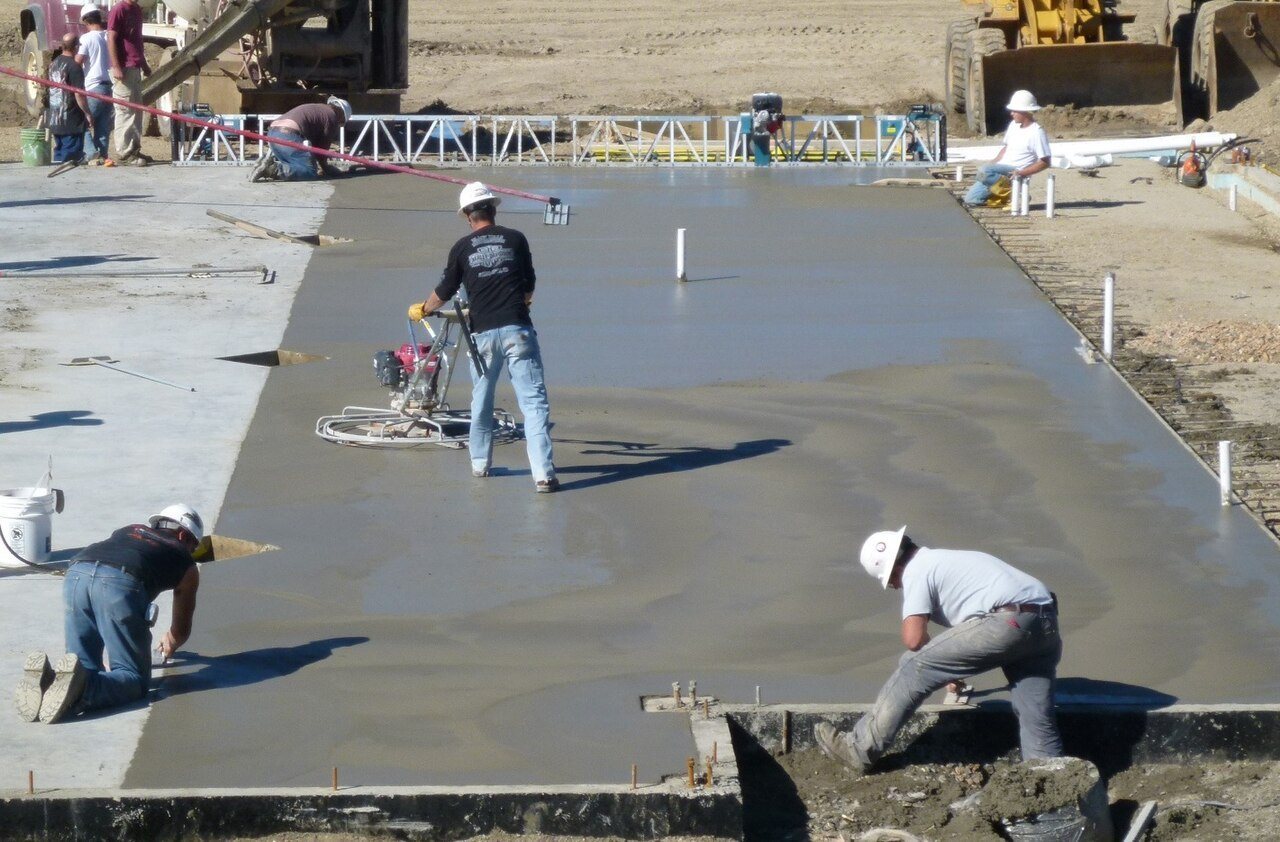 Crew finishing a large fresh concrete slab, one running a power trowel while others work the edges