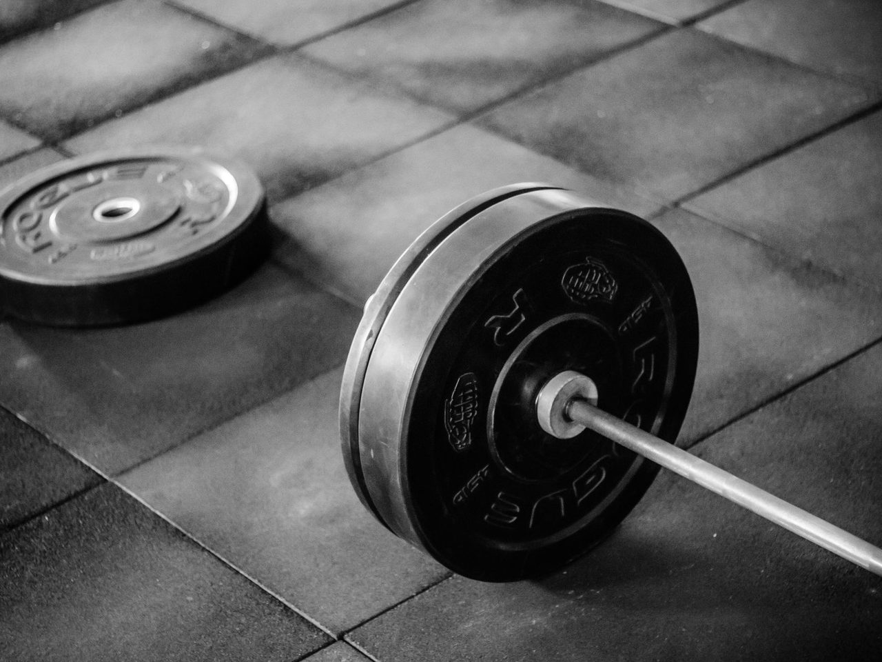 Grayscale photo of a barbell and weight plates on rubber floor tiles