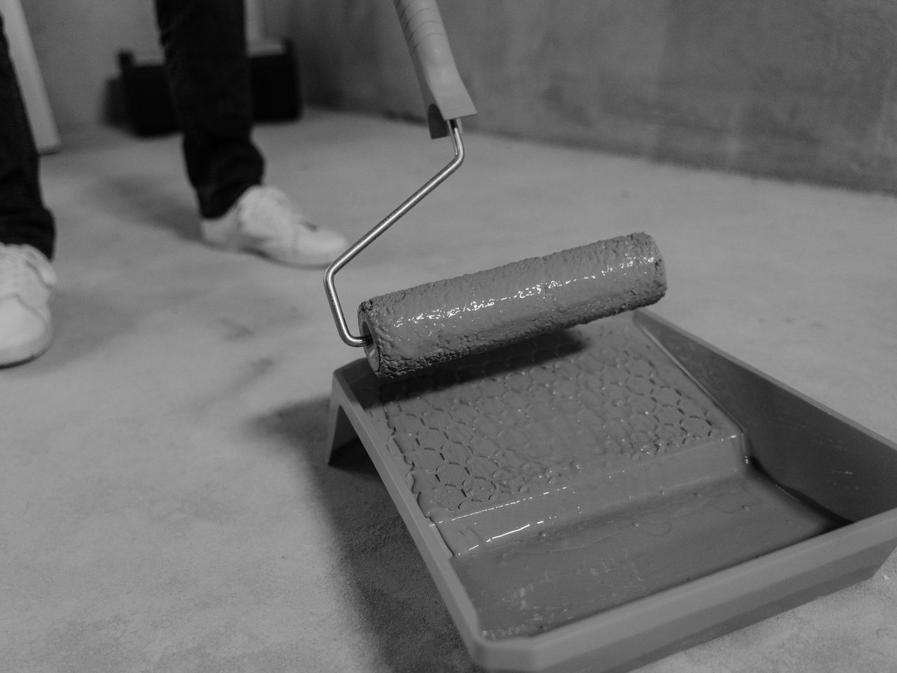 Black and white photo of a paint roller in a plastic tray on a concrete floor, with a person's legs and sneakers behind it.