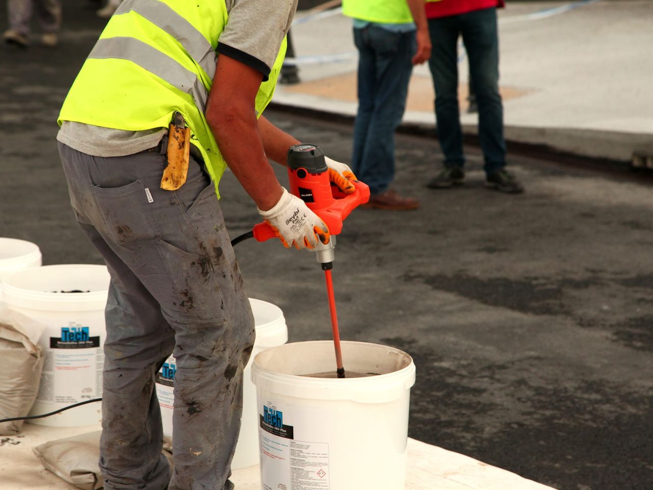 Worker in a yellow safety vest and gloves using a red electric paddle mixer in a white bucket, surrounded by bags and pails on a floor.
