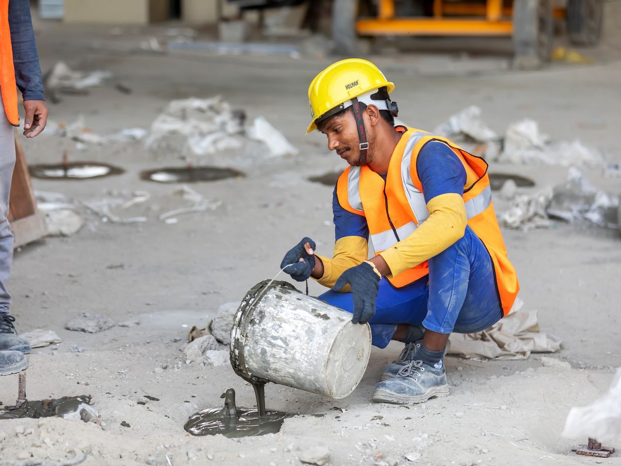 Crouching worker in a yellow hard hat, orange safety vest and gloves pouring gray liquid from a metal bucket onto a dusty floor.