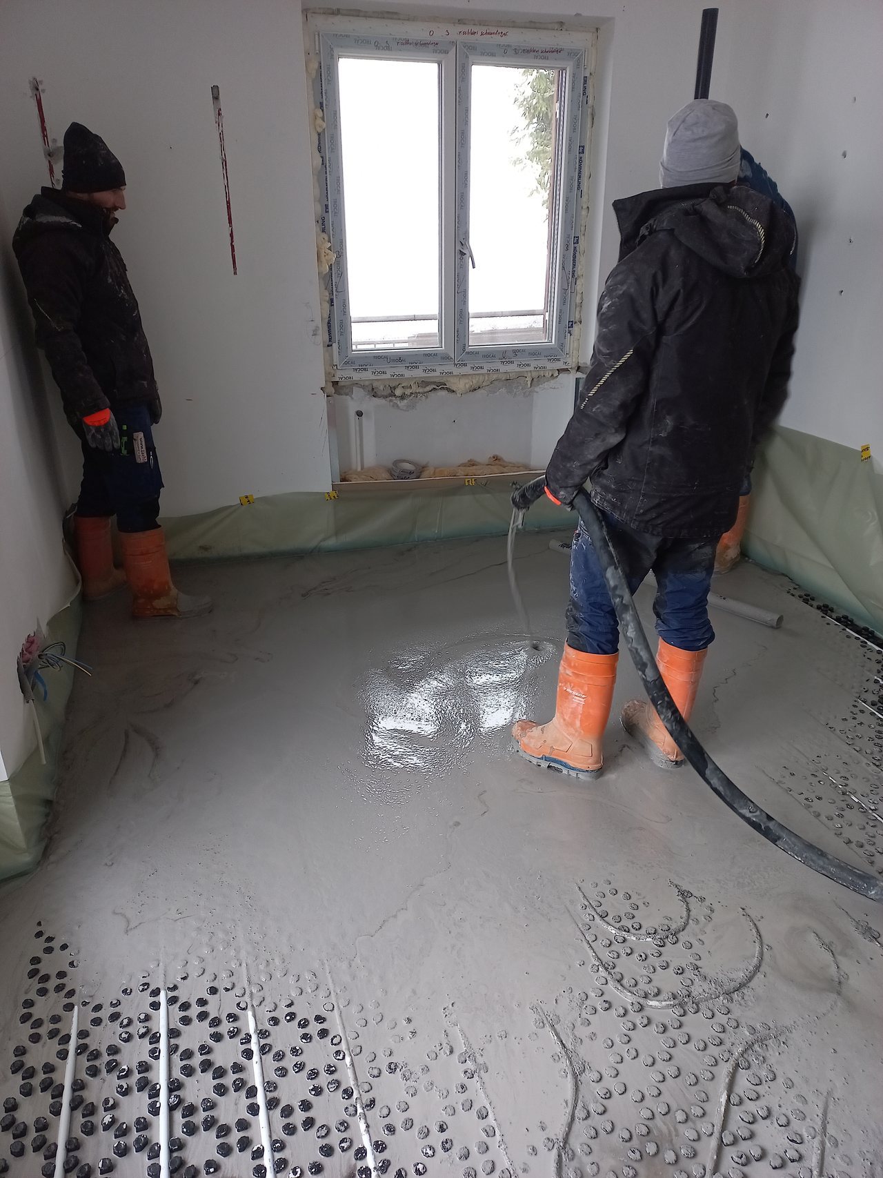 Two workers pumping liquid screed over underfloor heating panels in an unfinished room