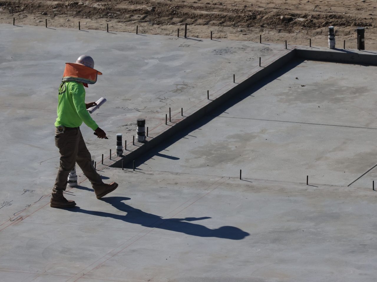 Worker in green jacket and orange hard hat walking on a large concrete slab with protruding pipes and dark edge forms in daylight.